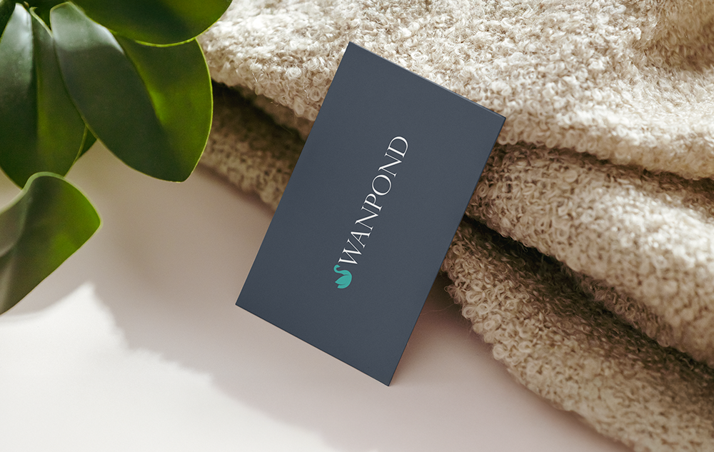 Swanpond eco resort logo design — dark navy business card with teal swan mark and white serif wordmark leaning against folded towels with a green plant