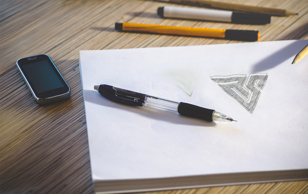 Early pencil sketch of the Revocare Solutions BPO company logo design showing the geometric RS triangle concept on white paper with pencils and pen on a wooden desk