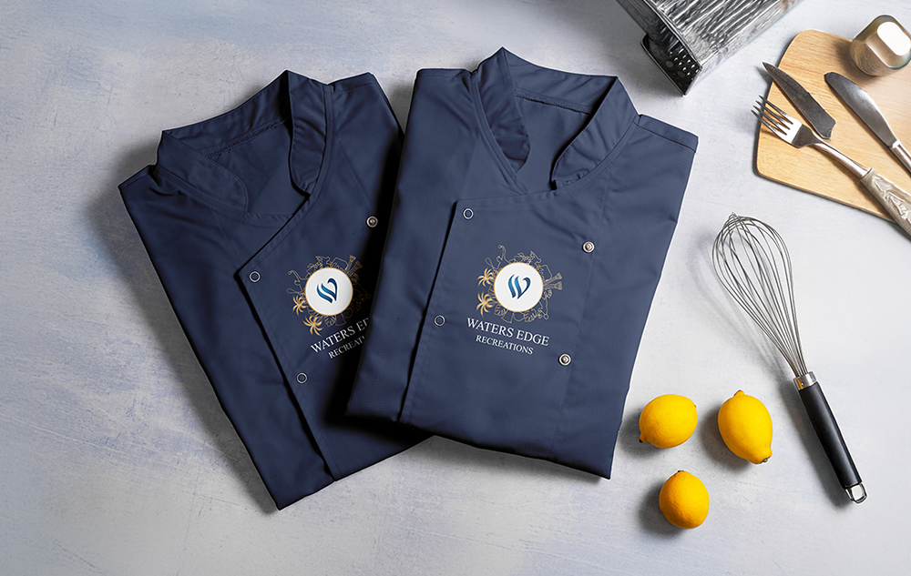 Waters Edge Recreations recreation logo design — two folded navy chef jackets with gold and blue emblem on chest pocket surrounded by lemons and a whisk on a light surface
