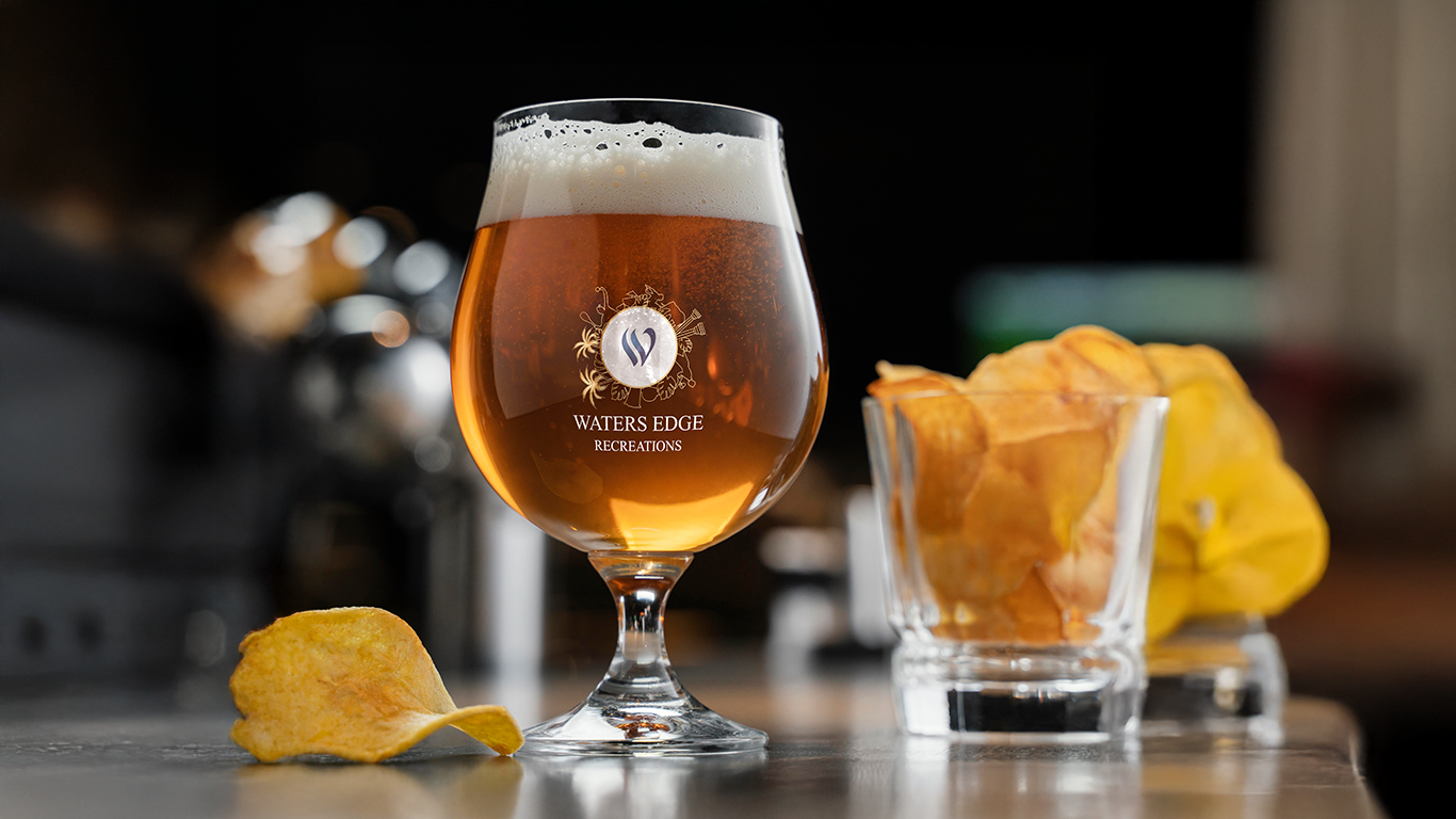 Waters Edge Recreations recreation logo design — branded tulip beer glass with gold emblem and white wordmark filled with amber beer on a bar counter with potato chips